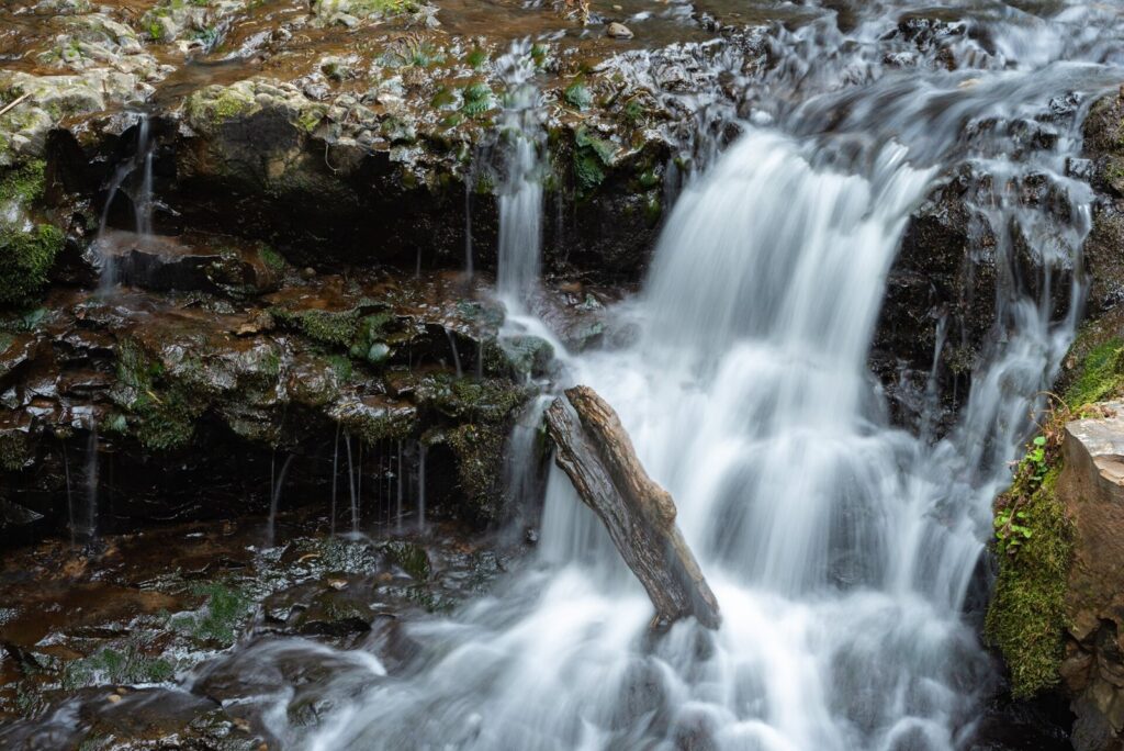 A small waterfall splashing over dark rock ledges into a pool, with a fallen log across the stream