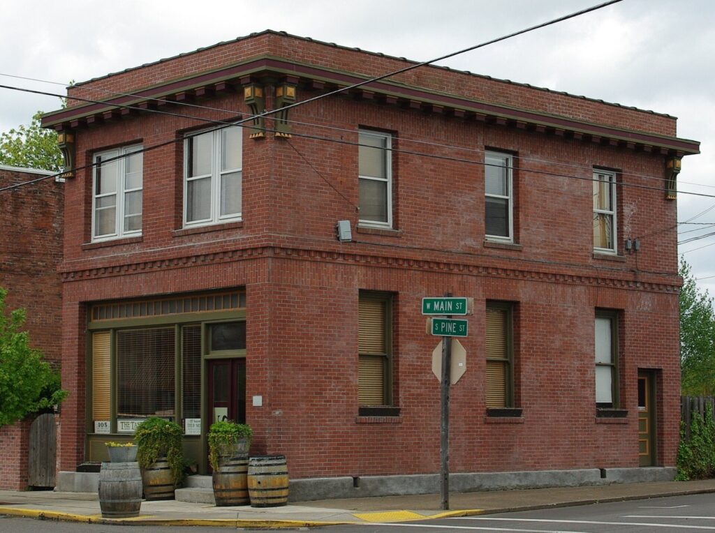 A two-story red brick commercial building on a small-town corner, with wine barrels and potted plants by the door