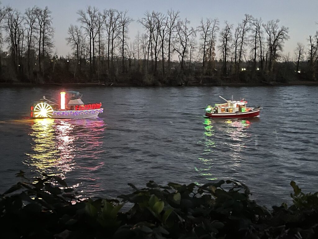 Two boats strung with colored holiday lights cruising a dark river at dusk, reflected in the water