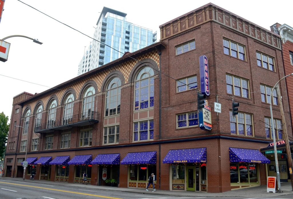 A three-story brick building with arched windows, blue starry awnings and a vertical Dance sign on a downtown street corner