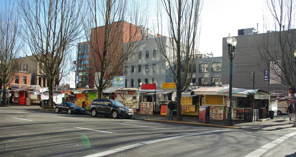 A row of food carts with awnings lining a downtown parking lot along a city street under bare trees