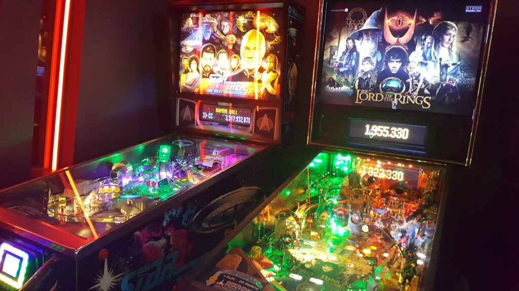Two brightly lit pinball machines side by side in a dark arcade