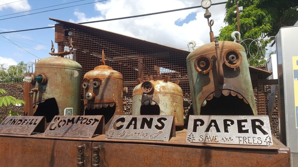 Four rusted metal sculptures with gaping mouths labeled landfill, compost, cans and paper serving as trash bins at Hawthorne Asylum