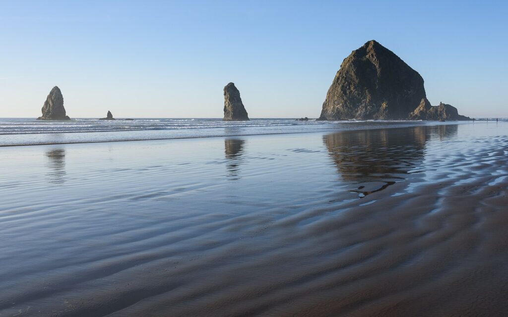 Haystack Rock and smaller sea stacks reflected in wet sand at low tide under a clear sky
