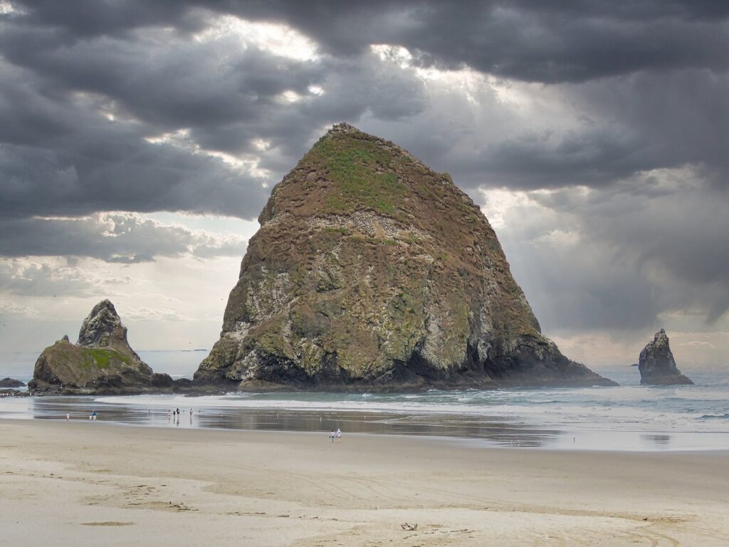 Haystack Rock under heavy storm clouds, with wet sand and tiny figures near its base at low tide
