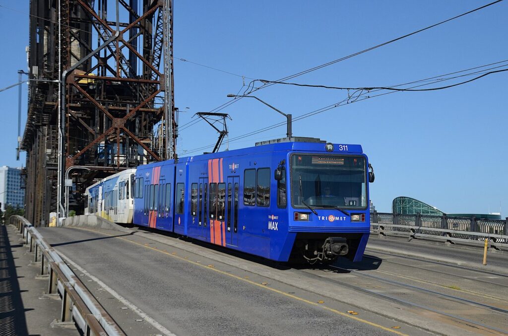 A blue TriMet MAX light rail train crossing the Steel Bridge under its steel lift tower on a sunny day