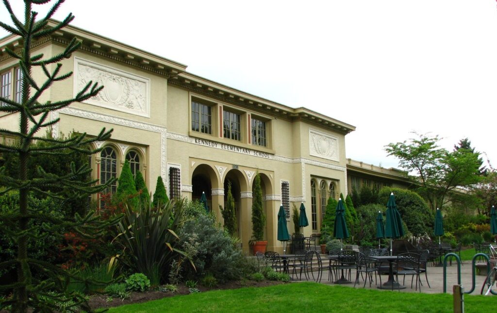 A pale stucco former school building with arched windows and a patio of umbrellas on a green lawn