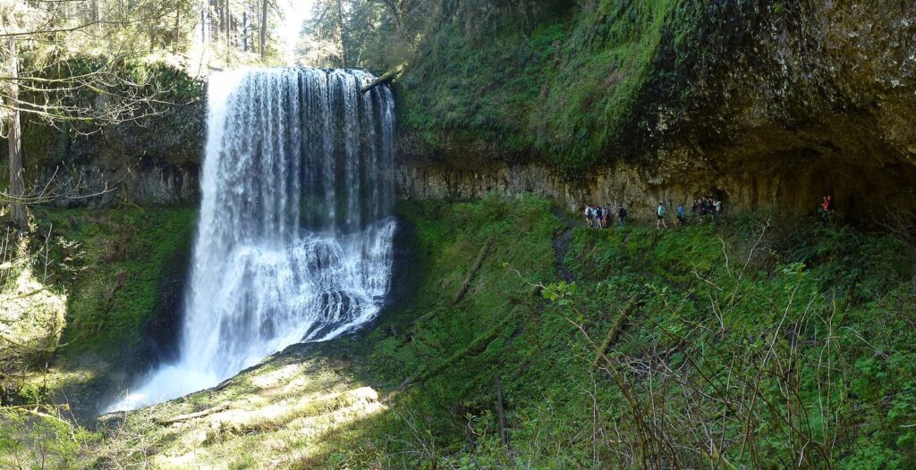 A wide waterfall pouring over a mossy basalt overhang, with a line of hikers on the trail that runs behind it