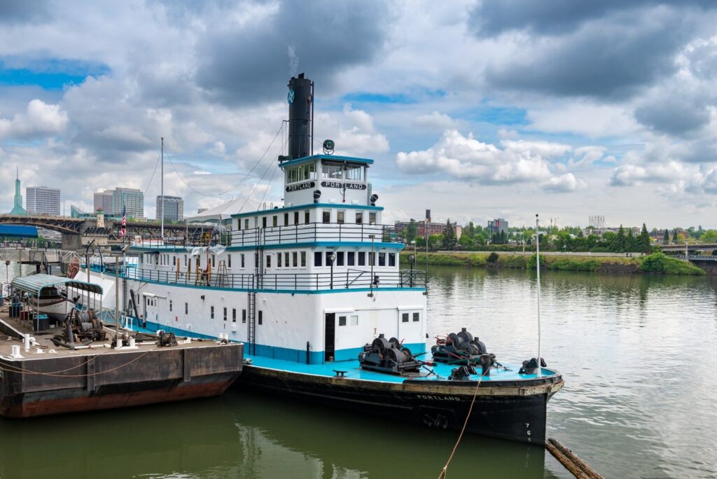 A white sternwheel steamboat with a tall black smokestack moored on a river beside a barge, with the city skyline beyond