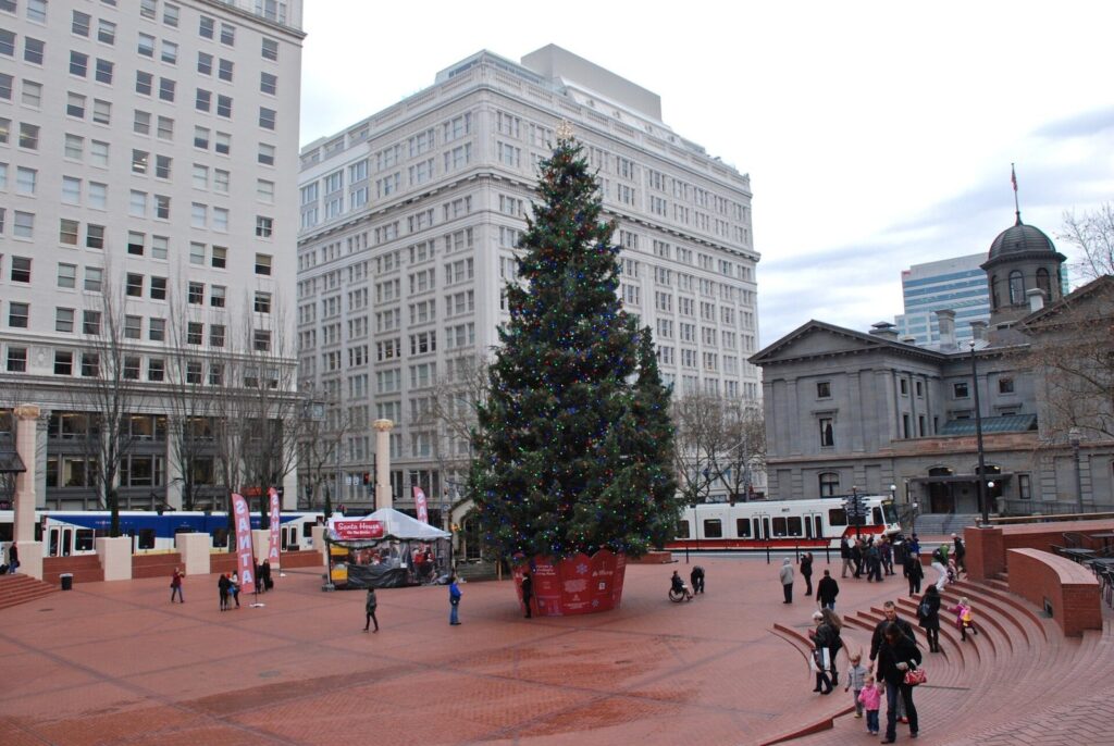 A decorated holiday tree in Pioneer Courthouse Square, with a MAX train passing in front of the old courthouse