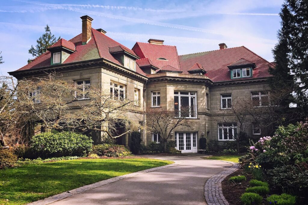 The sandstone walls and red tile roof of Pittock Mansion behind a curving drive and lawn