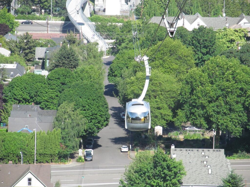 A silver Aerial Tram cabin hanging from its cables above treetops and a residential street