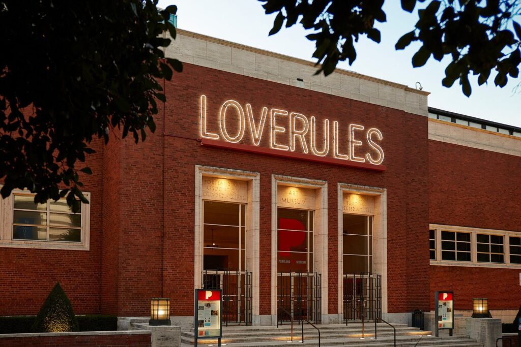 The brick facade and tall entrance doors of the Portland Art Museum at dusk, with a neon sign above the door