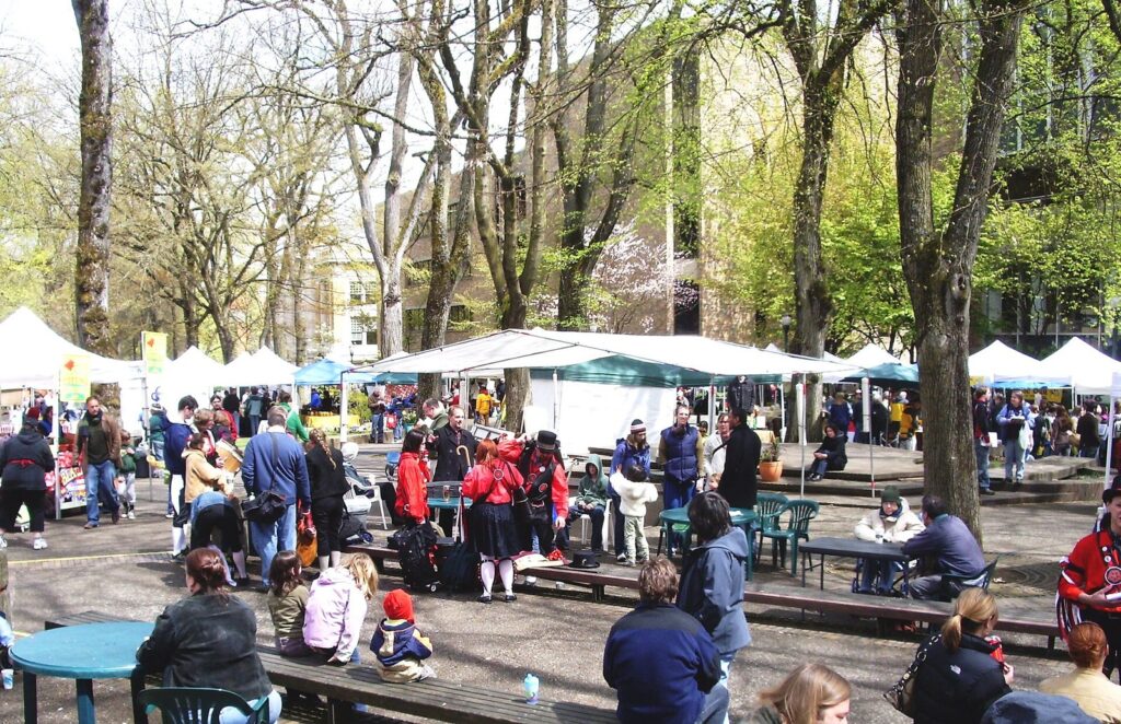 Shoppers and white vendor tents under tall trees in a leafy park on a spring day