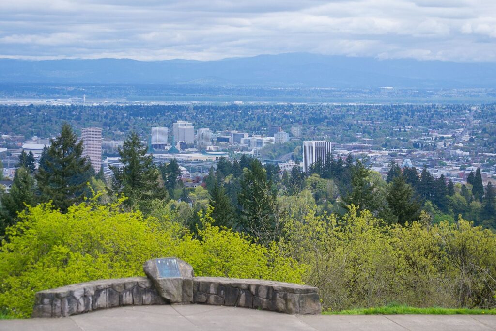 Downtown Portland's towers and the east side spread below a stone wall and spring foliage, with hills on the horizon