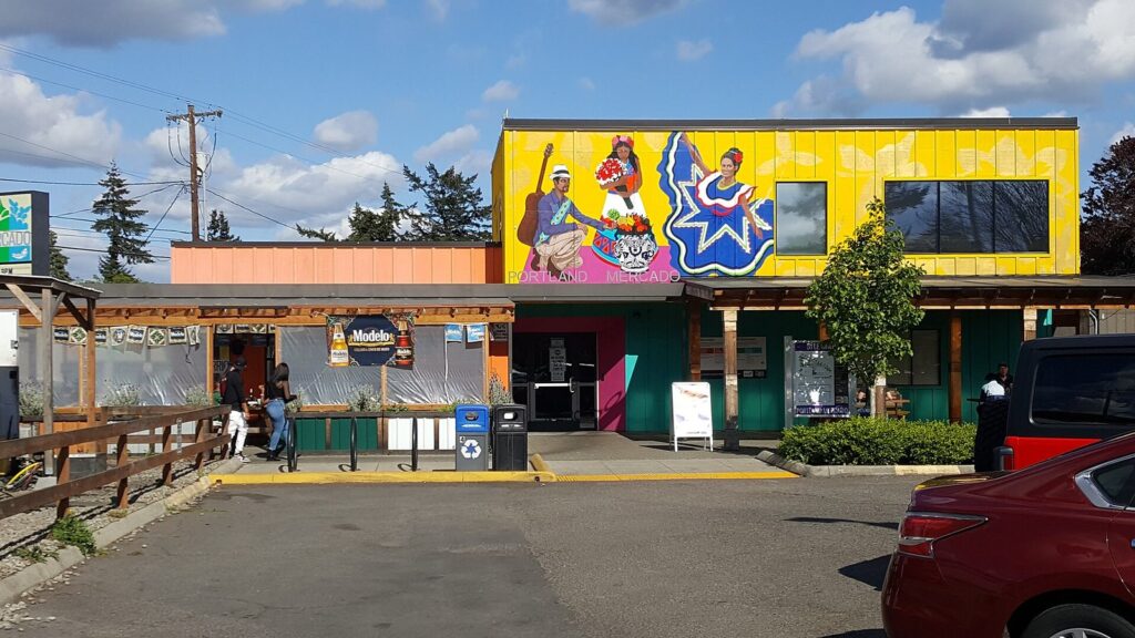 The Portland Mercado's colorful market building with a large folk-art mural above covered walkways and a parking lot