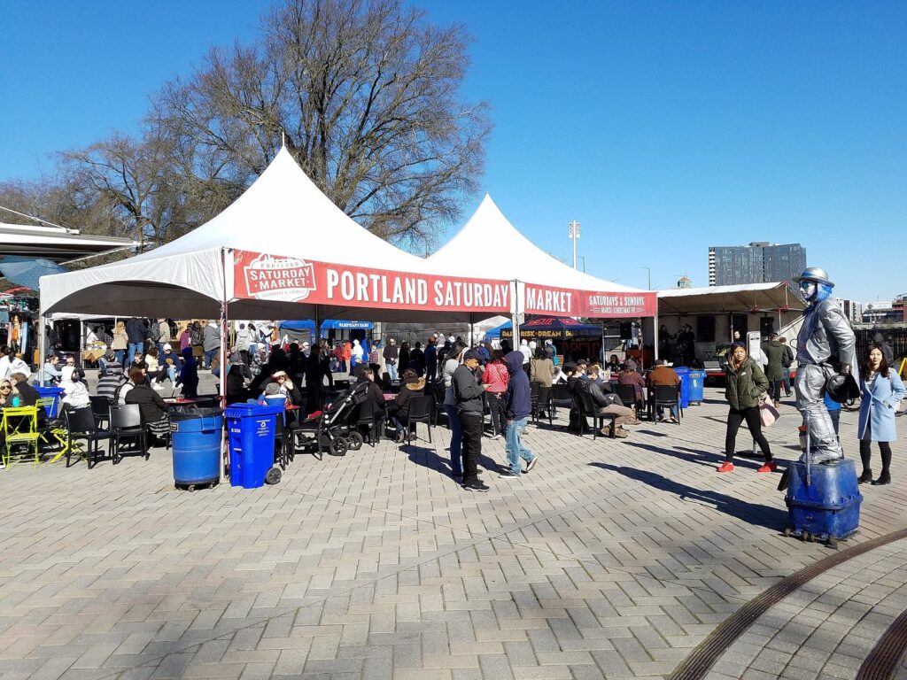 White market tents with a red Portland Saturday Market banner above a crowded brick plaza on a sunny day