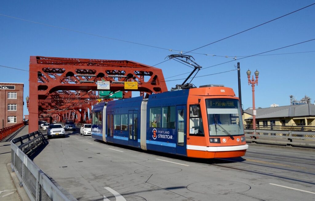 An orange and blue Portland Streetcar crossing the red steel trusses of the Broadway Bridge