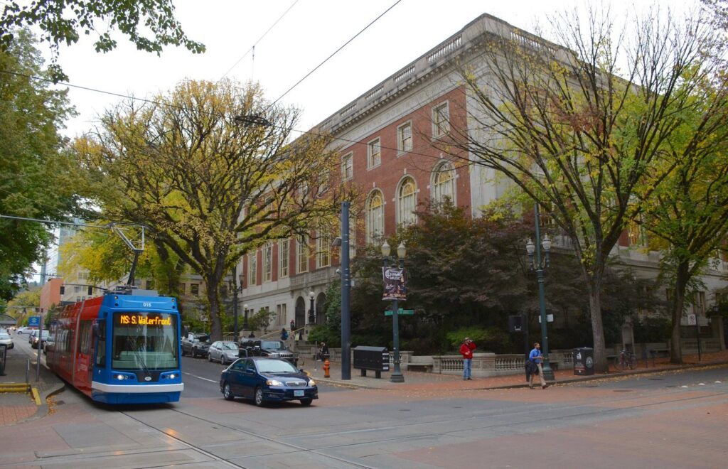 A red and blue Portland Streetcar signed for South Waterfront passing the brick Central Library under autumn trees