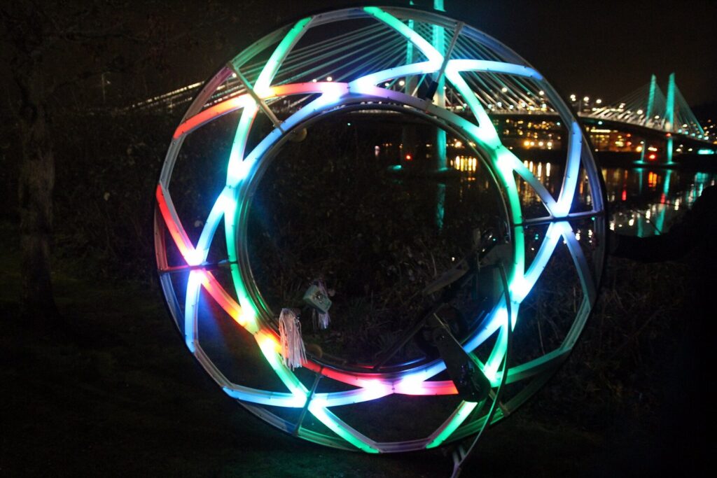 A glowing ring sculpture of colored lights on the riverbank at night, with Tilikum Crossing lit up across the water