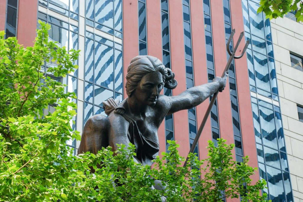 The dark hammered-copper Portlandia statue, a kneeling woman holding a trident, in front of a building's pink and glass facade