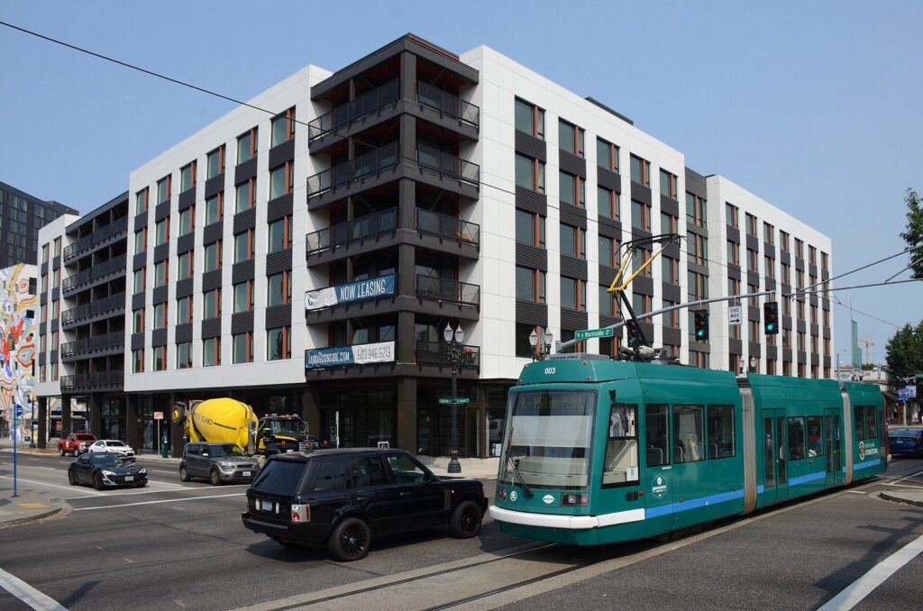 A teal Portland Streetcar on Grand Avenue passing a new apartment building at the corner of East Burnside