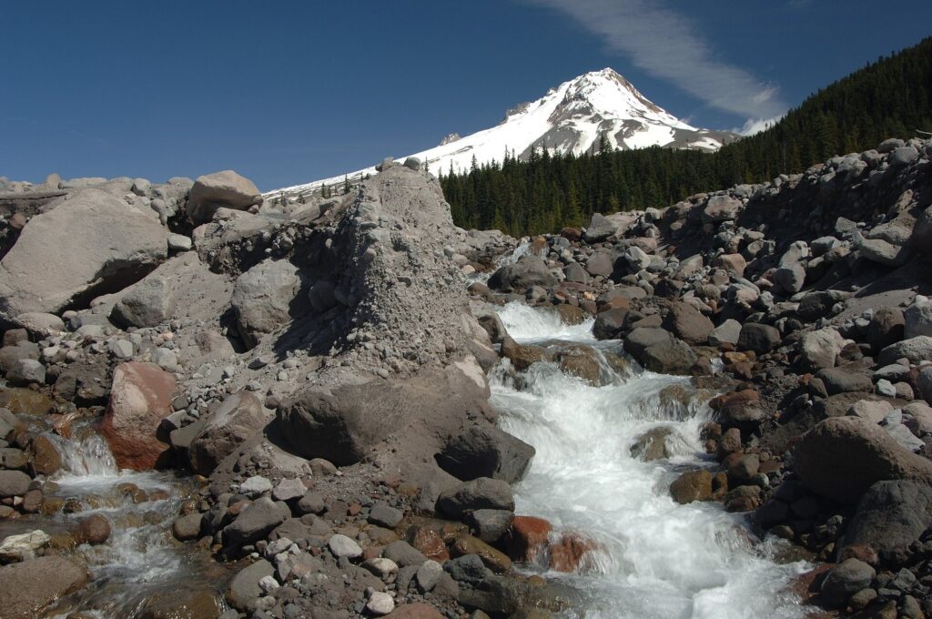 A rushing glacial stream among gray boulders, with a snowy volcanic peak and dark forest beyond
