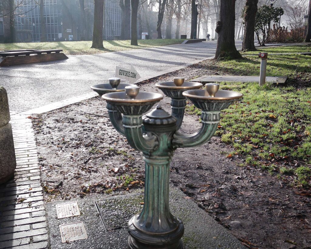 A bronze four-bowl drinking fountain on a brick sidewalk beside a lawn and trees on a misty morning