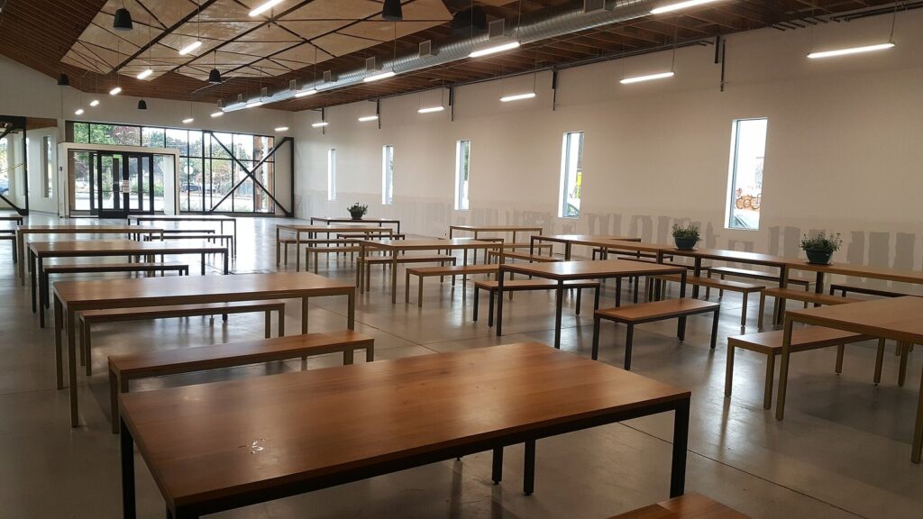 Long wooden tables and benches inside a bright food hall with tall windows and a high ceiling