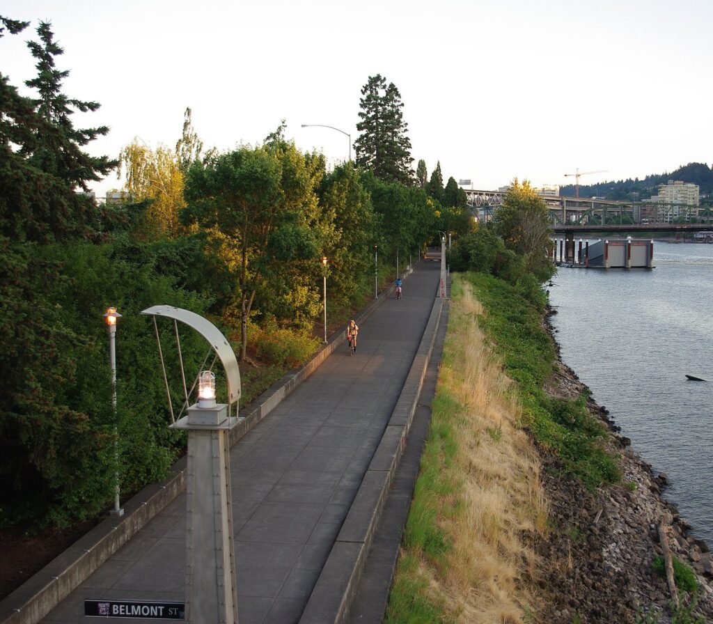 A paved riverside path with curved lamp posts and trees running beside the Willamette River, with a bridge in the distance