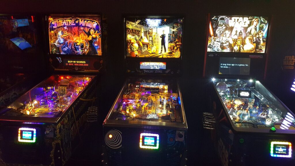 Three brightly lit pinball machines side by side in a dark arcade, their backglass art glowing