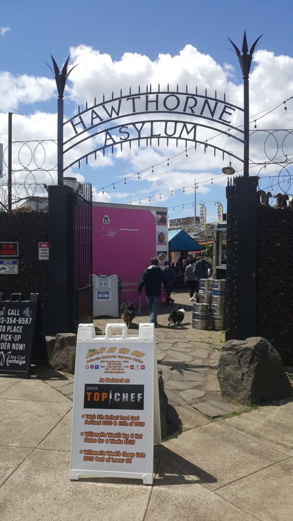 A wrought-iron arch spelling Hawthorne Asylum over the entrance to a food cart courtyard, with a sandwich-board sign in front