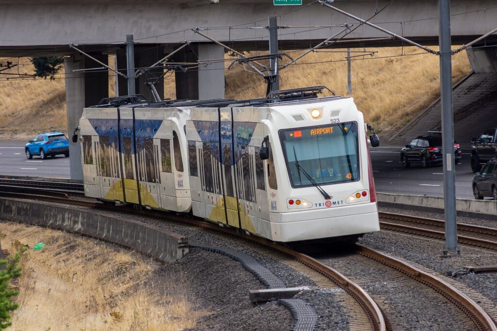 A white two-car MAX light rail train with an Airport destination sign curving along a track beside a highway overpass