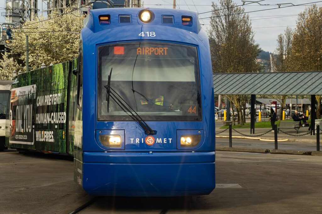 A blue MAX light rail train with an Airport destination sign pulling into a transit center platform under bare trees