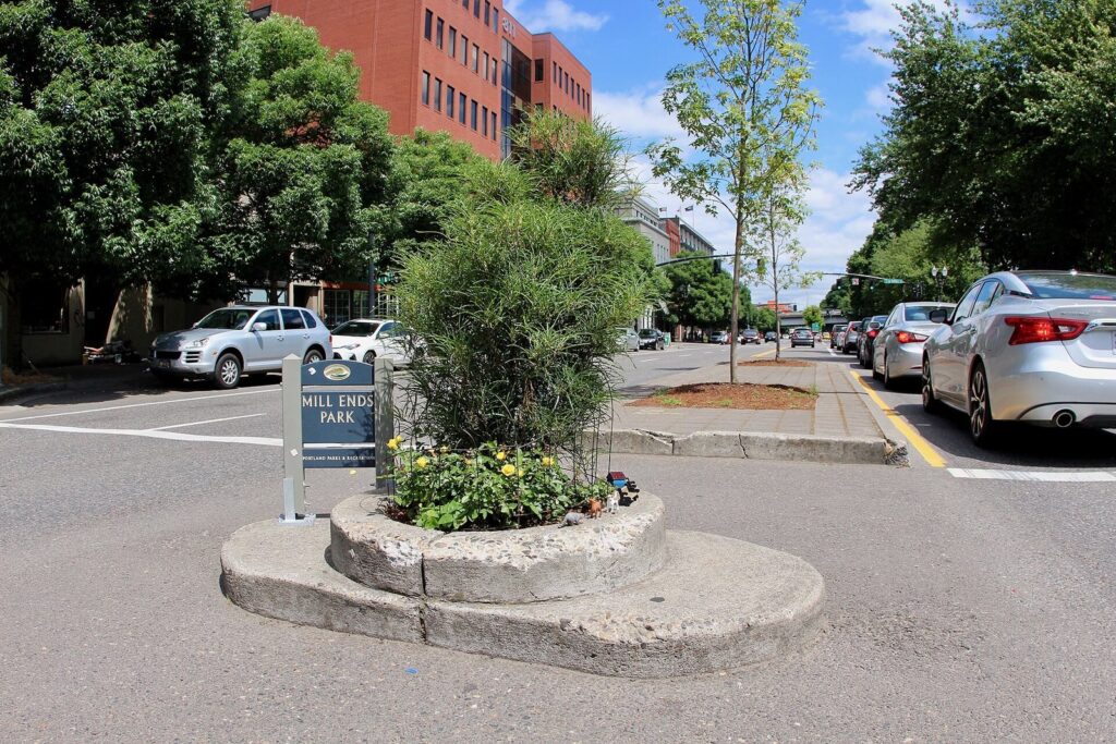 A tiny round planted park with a small tree and a sign in the median of a downtown street lined with parked cars