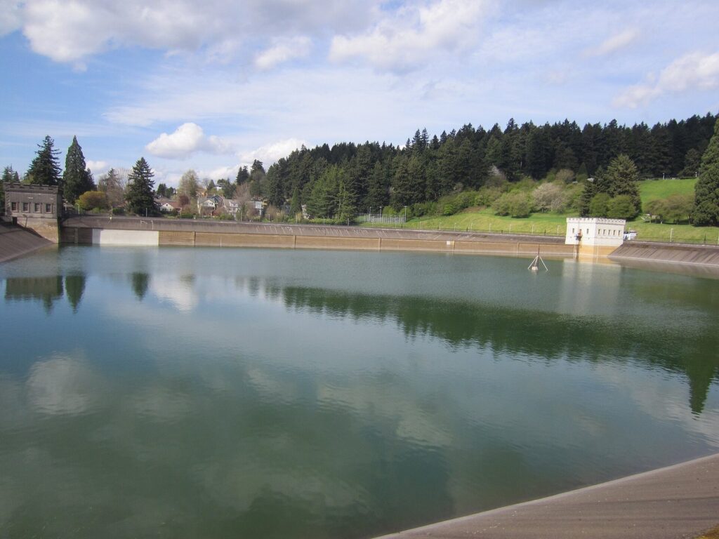 A wide open reservoir with a concrete rim and a small stone gatehouse, below a fir-covered hillside under a partly cloudy sky