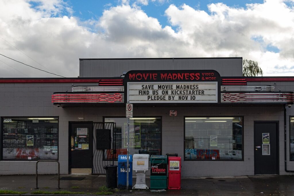 A low storefront with a red Movie Madness sign and a marquee asking for Kickstarter pledges under a cloudy sky