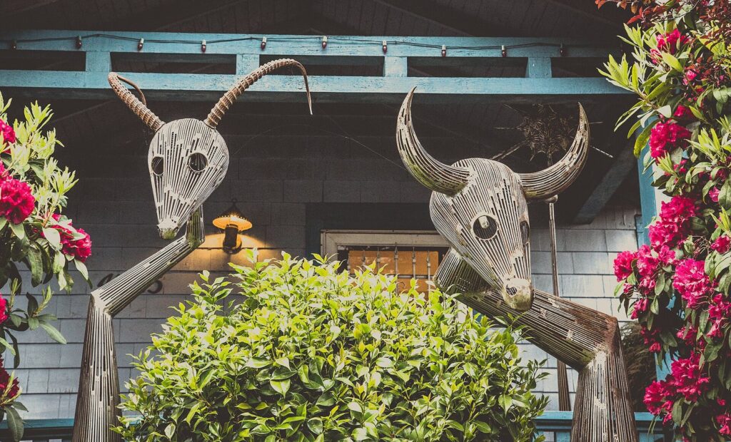 Two whimsical metal animal-head sculptures with long horns above a hedge, framed by flowering rhododendrons on a shop porch