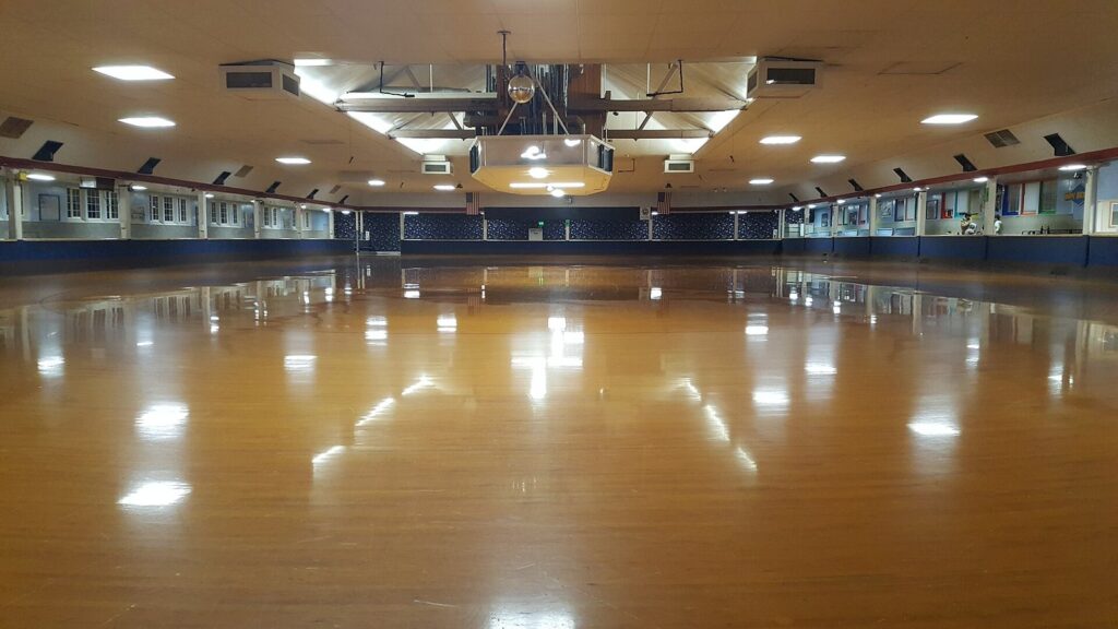 A wide, empty roller rink with a gleaming wooden floor under rows of ceiling lights, ringed by railings and windows