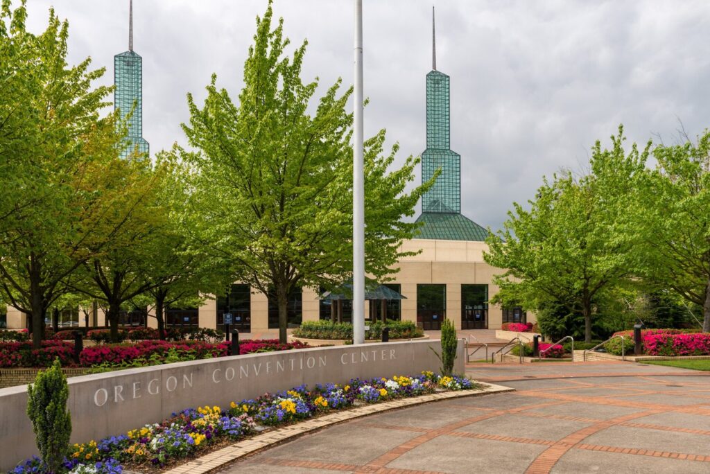 The twin glass spires of the Oregon Convention Center rising behind trees and a flower-lined plaza
