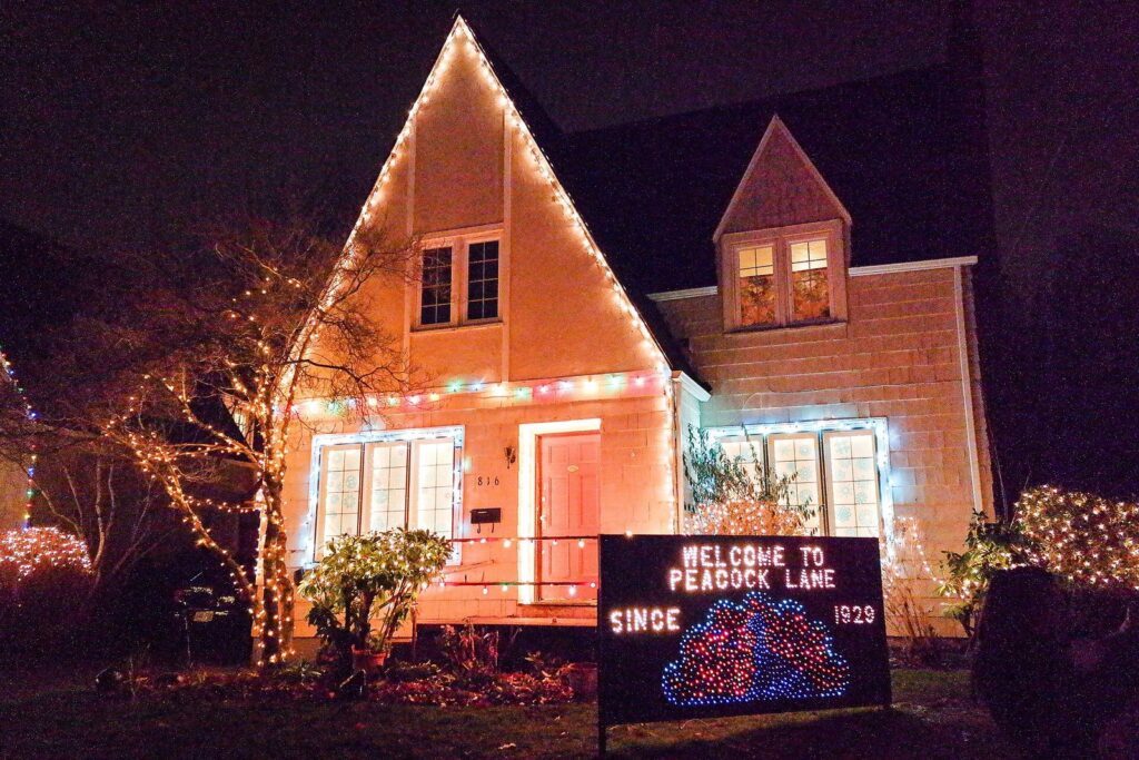 A steep-roofed house outlined in warm holiday lights at night, with a lit sign welcoming visitors to Peacock Lane