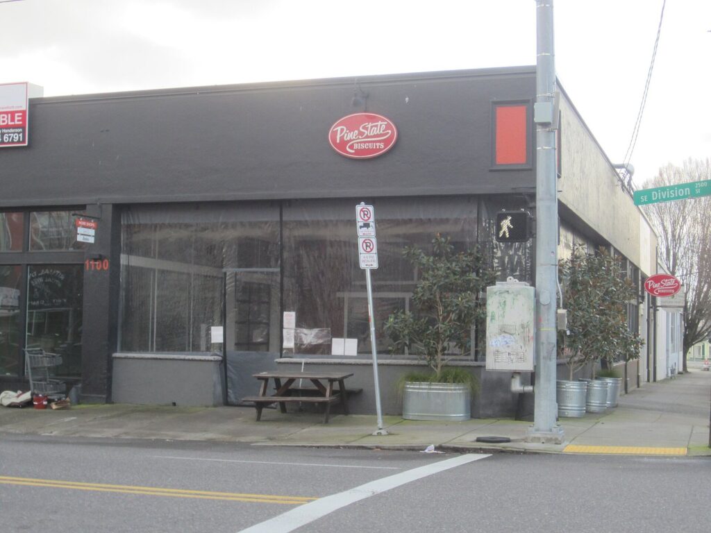A dark gray corner building with large windows, a red oval Pine State Biscuits sign and a picnic table on the sidewalk