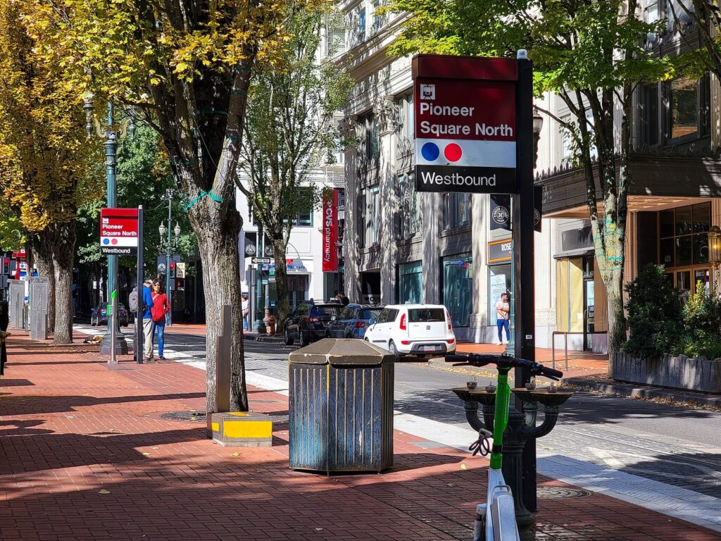 A red MAX station sign reading Pioneer Square North Westbound on a tree-lined downtown sidewalk with brick paving