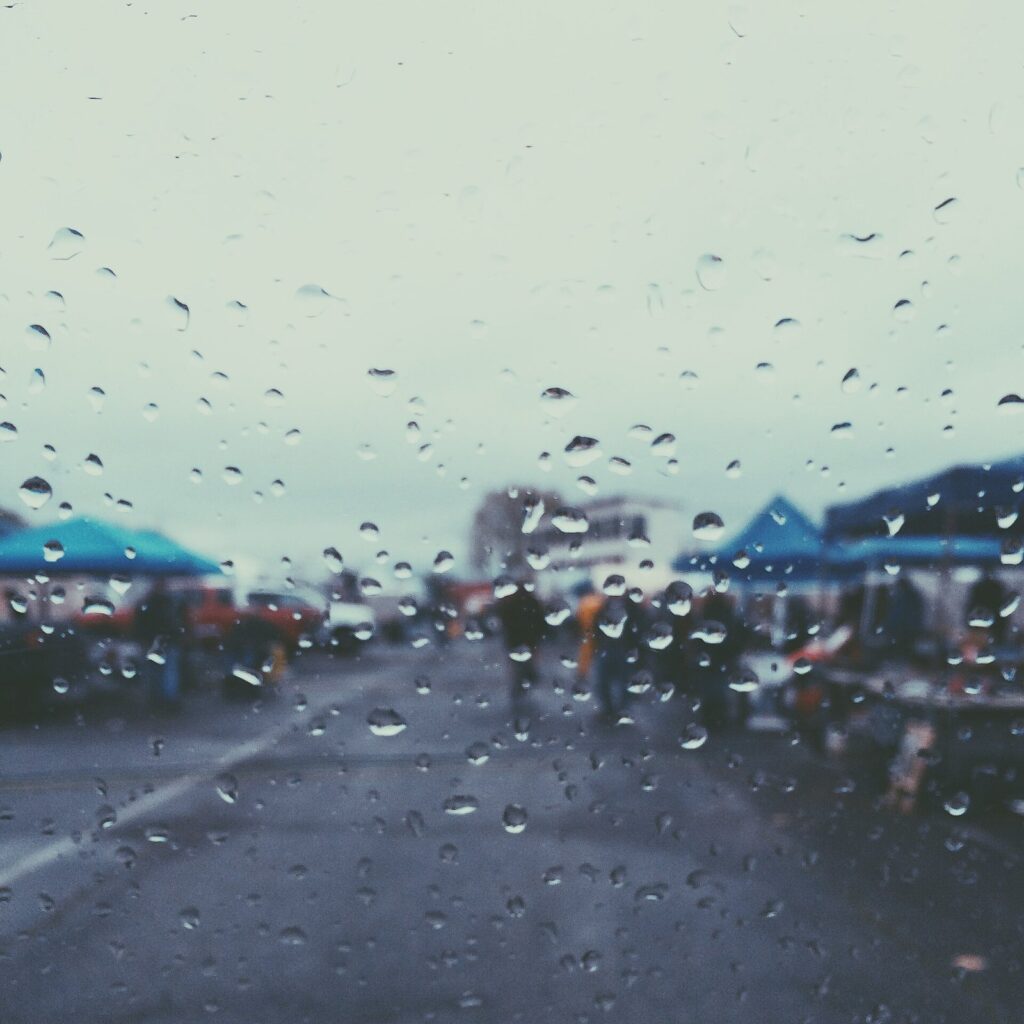 Raindrops on a car windshield blurring a street of blue market tents and people beyond