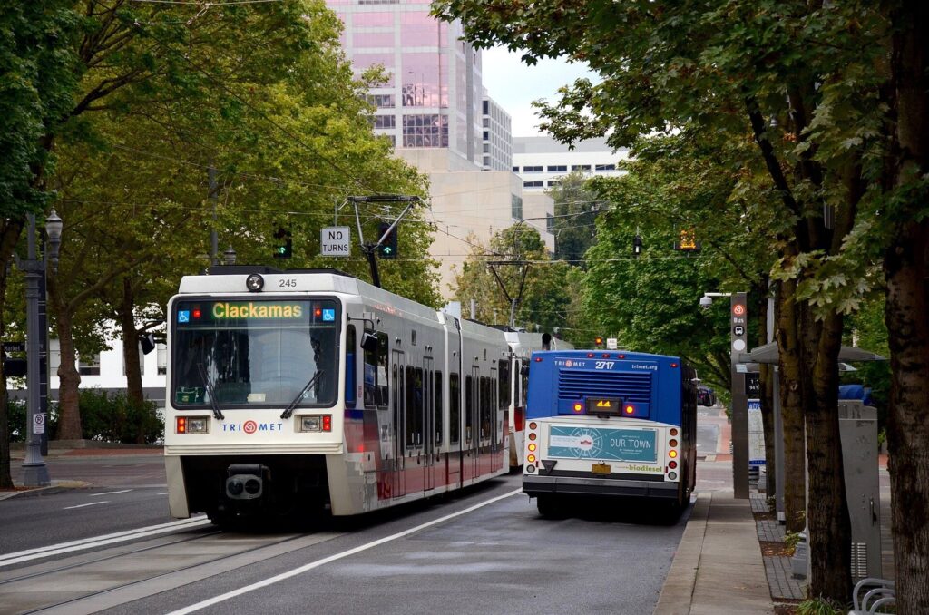 A white and red MAX light rail train and a blue TriMet bus side by side on a tree-lined downtown street