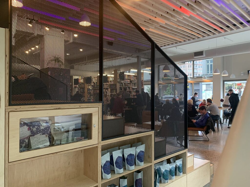 A bookstore coffee room with shelves, a perforated metal screen and readers at tables beside large windows