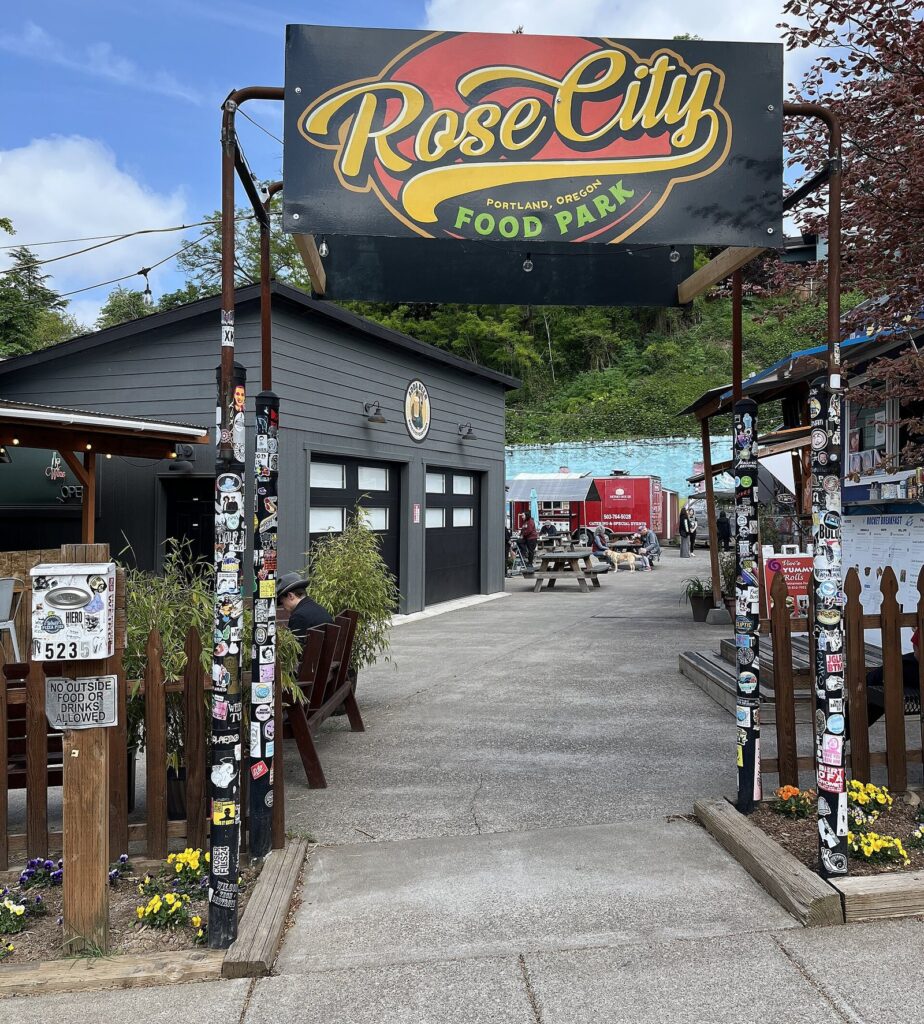 A Rose City Food Park sign over a paved entry path lined with sticker-covered posts, leading to carts and picnic tables