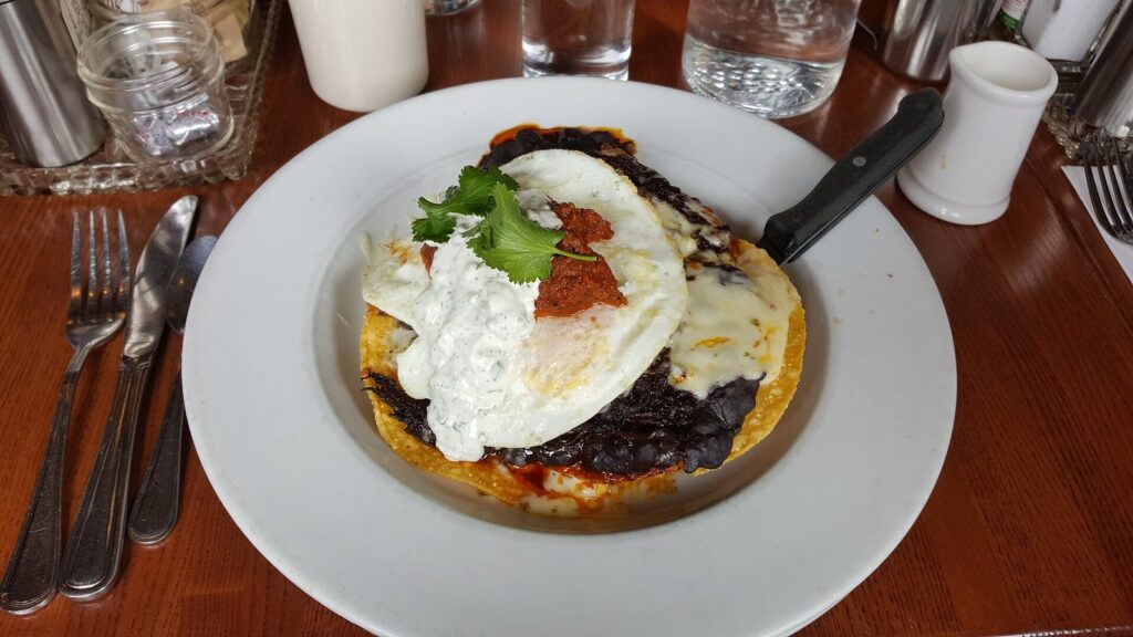 Huevos rancheros on a crisp tortilla with black beans, fried eggs, crema, red sauce and cilantro, on a white plate