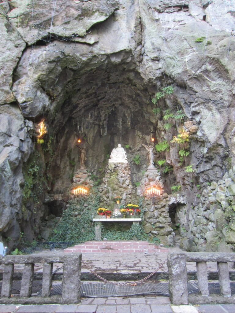 A white marble Pieta inside a rock cave at the foot of a cliff, lit by candles behind a low stone railing