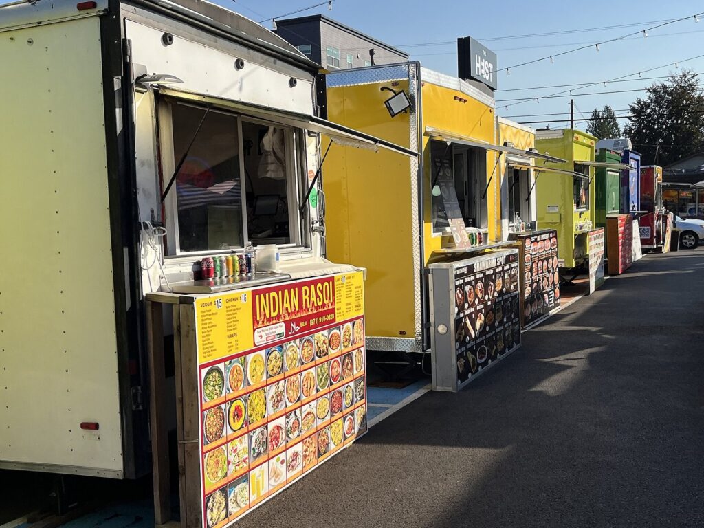 A row of brightly painted food trailers with menus on their counters lined up along a paved lot under string lights